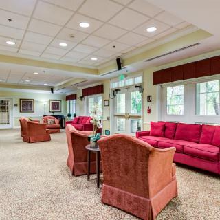 A spacious living room with comfortable seating, featuring red sofas and armchairs, carpeted flooring, large windows, and overhead lighting.