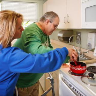 A caregiver assists an elderly person with cooking in the kitchen.