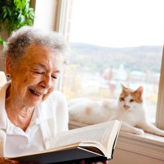 An elderly woman smiling while reading a book near a window with a cat lounging beside her.