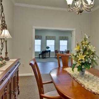 A dining room with a wooden table and chairs, featuring a floral centerpiece and a chandelier. In the background, there's a view of the living room with large windows and a piano.