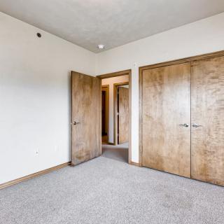 An empty bedroom with carpeted flooring, featuring wooden closet doors and a neutral color palette.
