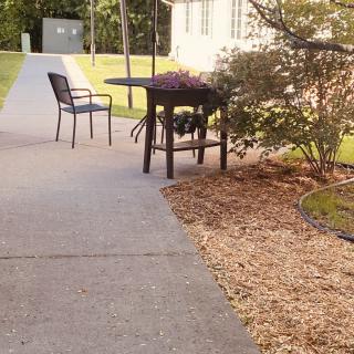 A patio set with a chair and table on a sidewalk surrounded by landscaped garden.