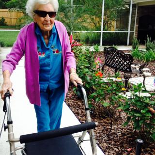 An elderly woman walking outside using a walker, enjoying the garden scenery.