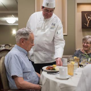 A chef serves a meal to an elderly couple at a restaurant.