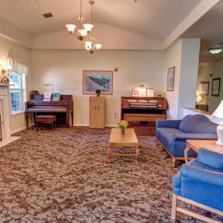 A cozy living room with blue sofas, a piano, and a chandelier, featuring a patterned carpet and wooden furniture.