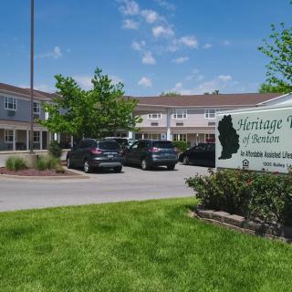 A residential building with a parking lot and sign, surrounded by greenery.
