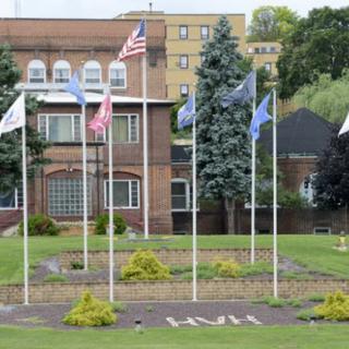 A row of flags in front of a brick building with surrounding greenery.