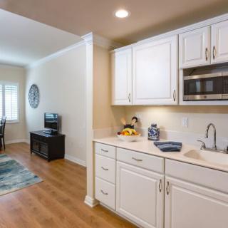 A modern apartment with a small kitchen and adjacent living area featuring white cabinets and wood flooring.