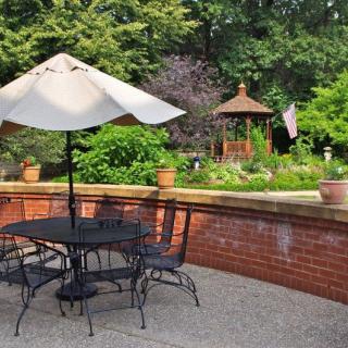 A tranquil patio setting with a shaded table and chairs, overlooking a lush garden with a gazebo.