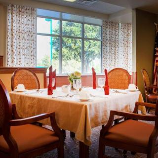 A dining room with a neatly set table, complete with red napkins and a flower centerpiece, next to a window with patterned curtains.