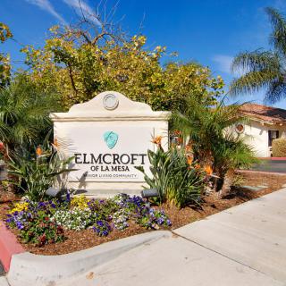 Entrance sign of Elmcroft of La Mesa surrounded by palm trees and flowers.