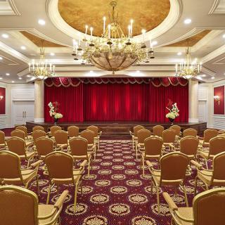 An elegant auditorium with red curtains and ornate chandeliers, featuring rows of comfortable seating.