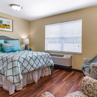 A cozy bedroom with a neatly made bed, featuring a patterned comforter, wooden flooring, and a window with blinds.