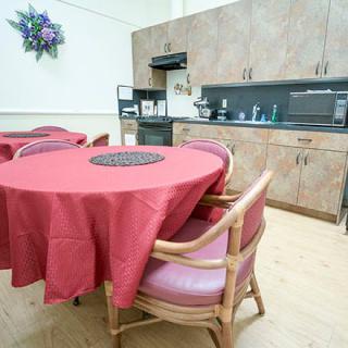 A kitchen and dining area with a round table covered in a red tablecloth, surrounded by chairs and wooden cabinetry in the background.