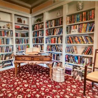 A cozy library with bookshelves filled with numerous books, a wooden table, and a patterned carpet.