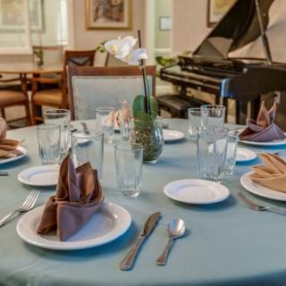 A dining table elegantly set with glasses, plates, silverware, and folded napkins in a room featuring a grand piano.