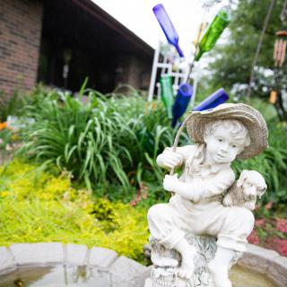 A decorative garden scene featuring a statue of a boy with a hat, surrounded by lush plants and colorful bottles.