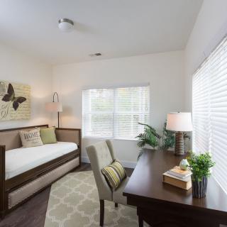 A cozy bedroom with a daybed, desk, and natural light from a large window.