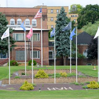 A neatly arranged row of flags flutters on poles in front of a brick building, with manicured lawns and trees adding to the scenic view.