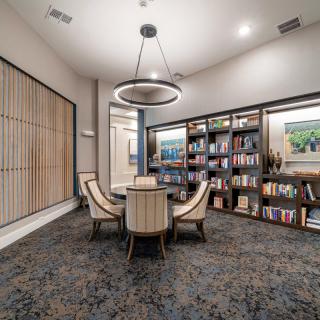 A modern library room with a round table, surrounded by upholstered chairs and bookshelves.