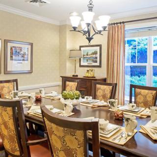 A well-decorated dining room with a large wooden table set for a meal, featuring elegant chairs, a chandelier, and natural light from a nearby window.