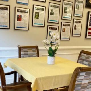 A dining area featuring a table with a yellow cloth, surrounded by chairs and adorned with a potted orchid, set against a wall decorated with framed articles.