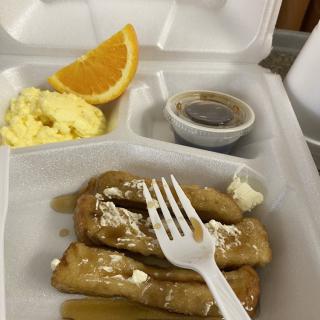 A takeout breakfast featuring pancakes with syrup, scrambled eggs, and an orange slice.
