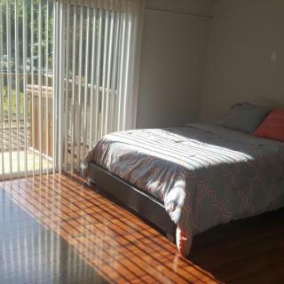 A sunlit bedroom with a bed and hardwood floors, featuring large windows with blinds.