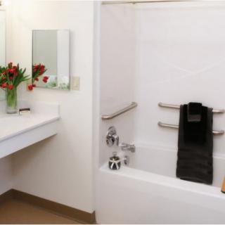A modern bathroom featuring a bathtub with grab bars, a neatly arranged towel, a large mirror above the sink, and a vase of red flowers.