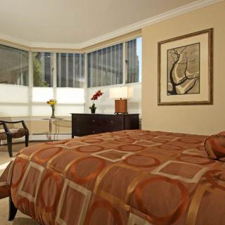 A modern bedroom with a large bed, geometric-patterned bedspread, and contemporary furniture, featuring an artwork on the wall and a corner window allowing natural light.