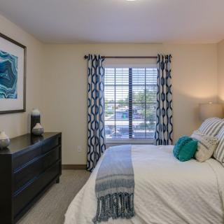 A cozy bedroom with a neatly made bed, modern dresser, and patterned curtains framing the window.