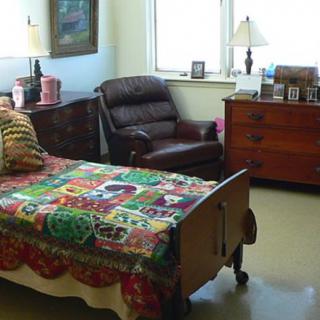 A cozy bedroom featuring a bed with a colorful quilt, an armchair, and wooden furniture including a dresser and a lamp.