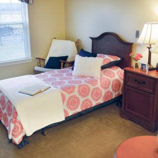 A cozy bedroom with a single bed, featuring a floral bedspread, a wooden nightstand with a lamp, and an armchair by the window.