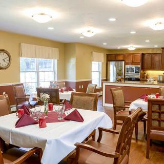 A well-lit dining room with neatly arranged tables and chairs, adjacent to a kitchen area.