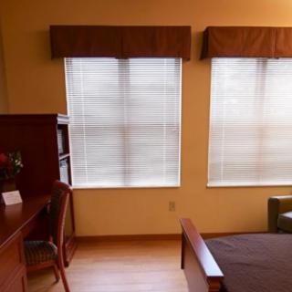 A cozy bedroom with a desk, chair, and large windows with curtains.