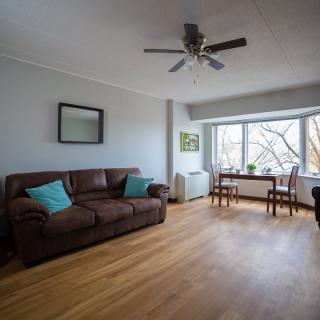 A bright and minimalist living room with a large window, featuring a brown leather sofa, hardwood floors, and a ceiling fan.