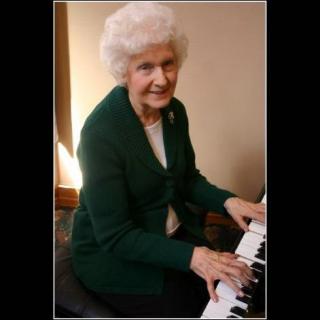 An elderly woman playing the piano, dressed in a green jacket and smiling warmly.