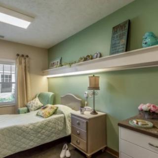 A cozy bedroom with pastel green walls, featuring a single bed, a nightstand with a lamp, and decorative items on shelves.