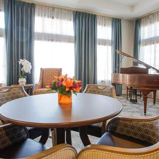 A cozy living room featuring a round table with a floral centerpiece, cushioned chairs, and a grand piano near large windows with curtains.