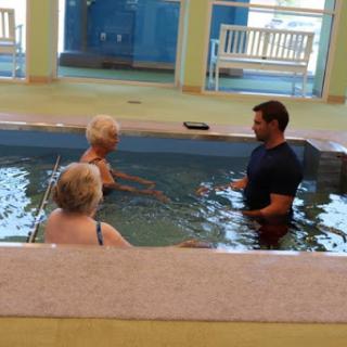A water therapy session taking place in a swimming pool with a trainer assisting two adults.