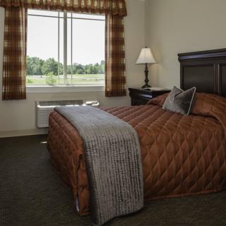 A cozy hotel bedroom with a brown quilted bedspread, a bedside lamp, and large window with plaid curtains.