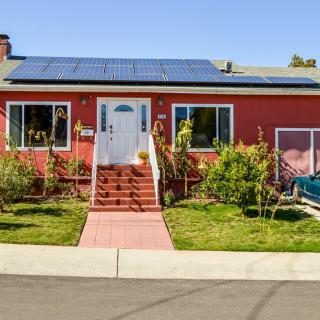 A red house with solar panels on the roof, featuring a front yard garden and steps leading to the entrance.
