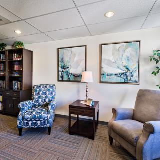 A cozy office interior featuring two chairs, a wooden bookshelf, artwork on the wall, and a small lamp on a side table.