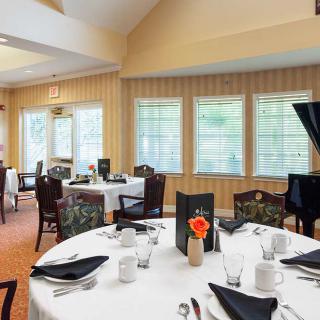 An elegant dining room with tables set for service, featuring white tablecloths, black napkins, and a grand piano by the window.