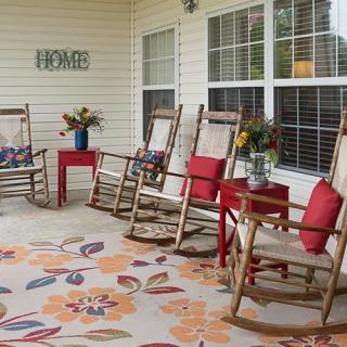 A cozy porch with wooden rocking chairs, red cushions, a floral rug, and vibrant flower arrangements.