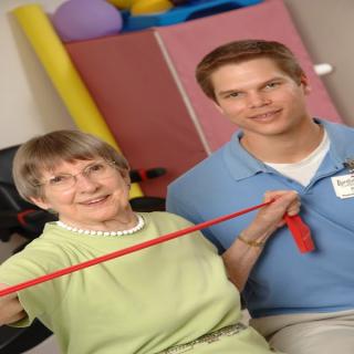 An elderly woman participating in a physical therapy session with a therapist, using a resistance band for exercise.