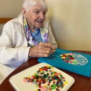 An elderly woman engaging in crafting with colorful buttons on a table.