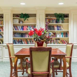 A cozy library room with a wooden table, chairs, and bookshelves adorned with colorful books and a floral centerpiece.