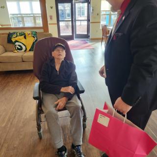 A man in a wheelchair is being visited by another person holding a gift bag indoors.