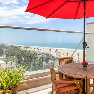 A scenic beach view from a balcony featuring wooden patio furniture and a red umbrella.